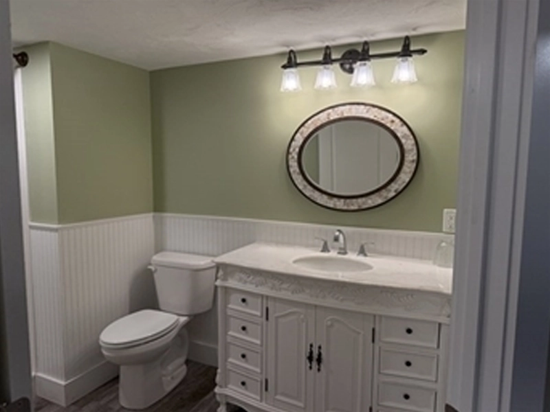 A bathroom with a white vanity, oval mirror, toilet, and green walls with white paneling.