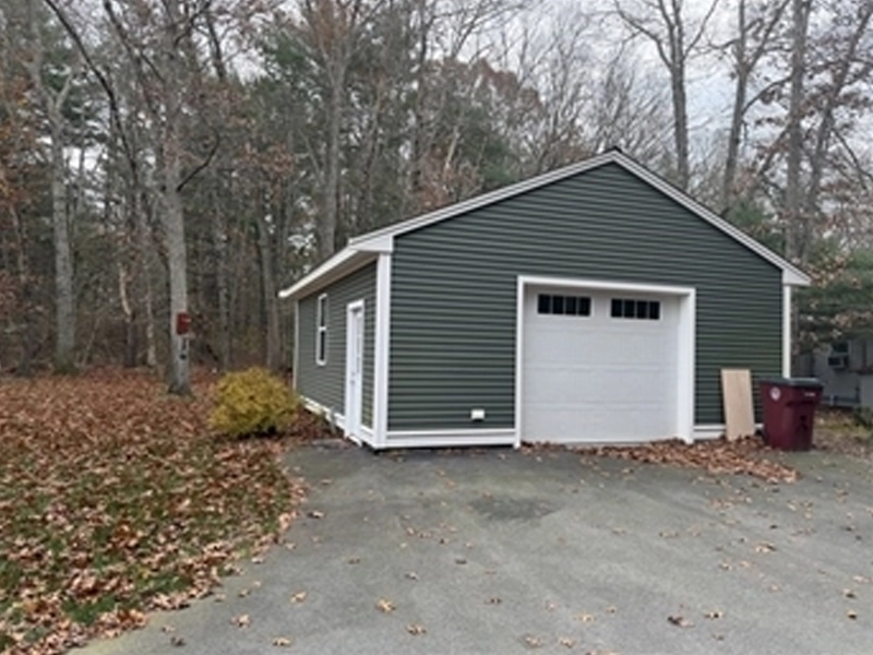 Green garage with white trim, surrounded by trees and fallen leaves on a gray driveway.