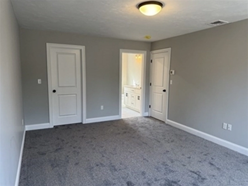 Empty gray-carpeted bedroom with two doors and a doorway leading to a lit bathroom.
