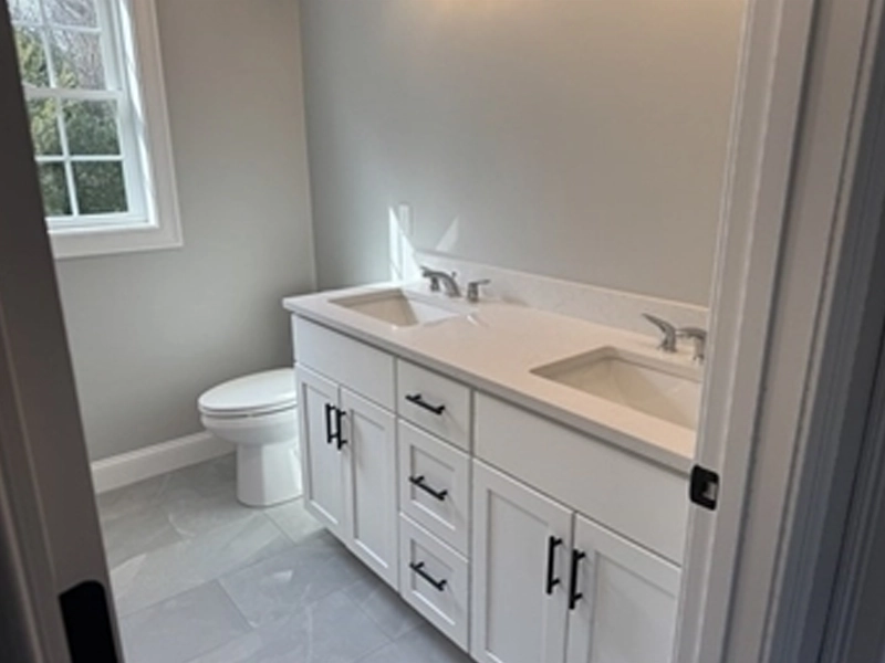 White bathroom with double sink vanity, toilet, and a window letting in natural light.