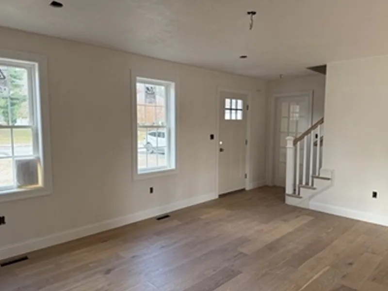 Empty room with light wood floors, white walls, two windows, and a door near a staircase.
