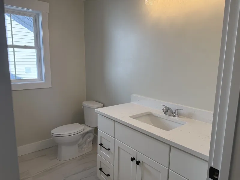 Modern bathroom with white vanity, sink, toilet, and window on light-colored walls.