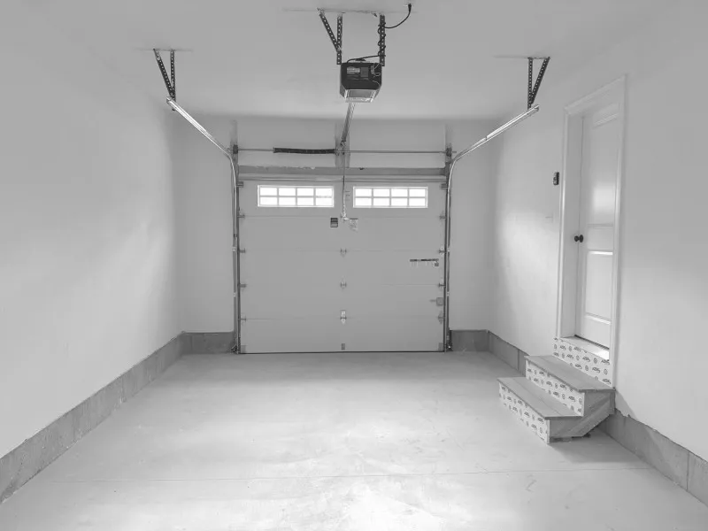 Empty garage with closed door, overhead opener, and steps leading to a side door on the right.