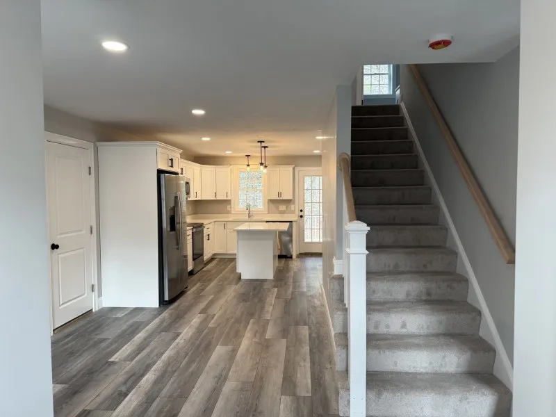 Modern kitchen with white cabinets, island, and stairs with gray carpeting on the right.