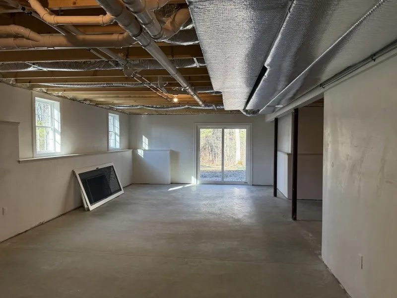 Unfinished basement with exposed pipes, concrete floor, and sunlight from windows and door.