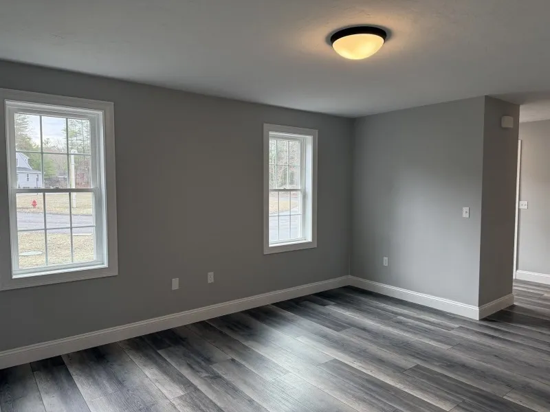 Empty room with gray walls, two windows, and dark wood flooring. Ceiling light is on.