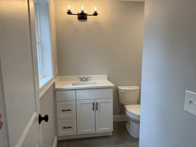 Small bathroom with white vanity, toilet, wall light fixture, and neutral gray walls.
